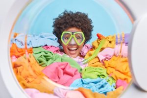Cheerful curly haired young busy woman wears snorkeling mask for diving has lot of laundry to make does housework drowned in multicolored clothes against blue background. Housekeeping concept